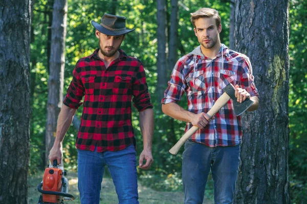 Two friends Lumberjack worker in the forest. Woodcutter with axe and lumberjack with chainsaw. Handsome woodcutter men hipsters in forest