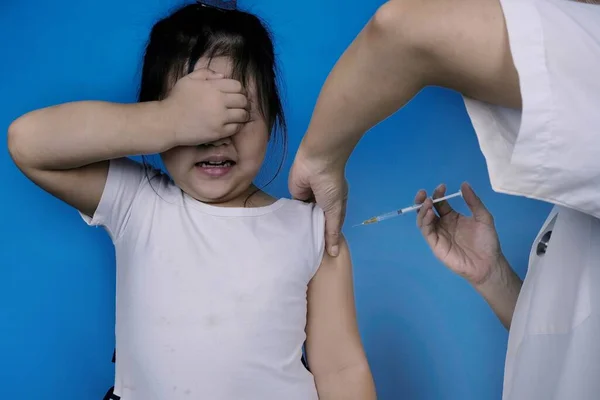 小孩在医院被注射吓坏了 女孩在儿科诊所接种流感疫苗时害怕针头盖住脸 蓝色背景 免版税图库照片
