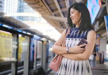 Beyaz ve mavi elbiseli Asyalı bir iş kadını kollarını kavuşturmuş istasyonda sabah işe gitmek için hava trenini bekliyor..