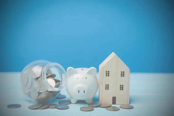 Konsept, krediyle geleceğe ev, mülk, tasarruf, mülk satın almak için tasarruf sağlar. Bardağında bozuk paralar olan beyaz bir kumbara ve eski ahşap masa mavi arka planda kopya alanı olan ahşap ev modelleri..