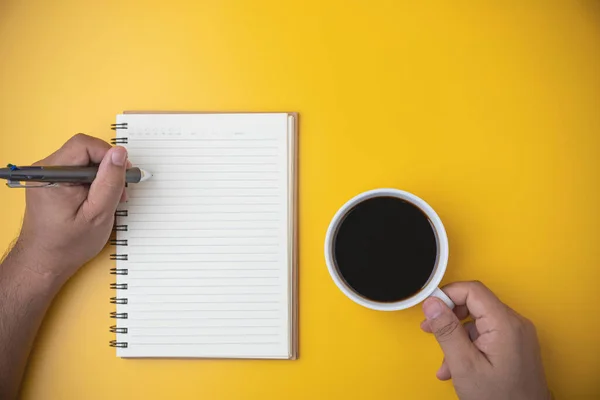 Morning drinks coffee. top view espresso coffee break. Hand of a man holding a white cup with a hot black coffee on yellow background with copy space. A man uses a pen to write a diary in notebook.