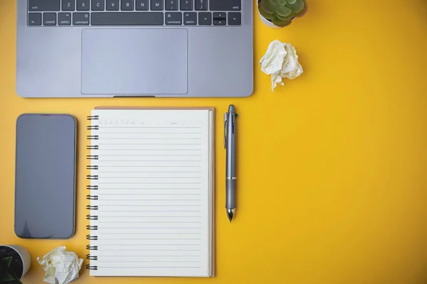 Flat lay of office yellow desk with laptop, and smartphone. Top view blank notebook with a pen on yellow table with copy space. Business, technology, study, and learning concept.