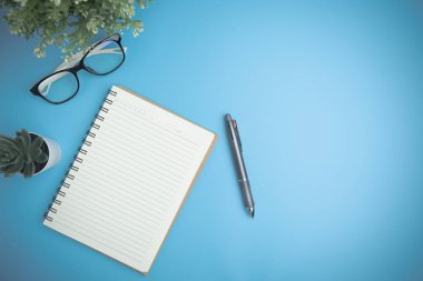 The blue office desk with a notebook, a pen, glasses, and a mini tree on the blue desk flat lay, background. Top view workspace of table office with copy space. Business, study, learning concept.
