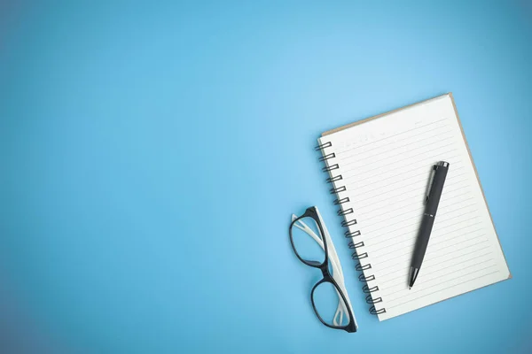 The blue office desk with a notebook, a pen, and glasses flat lay. Office table desk of the business flat lay, background with copy space. Top view workspace of table office.