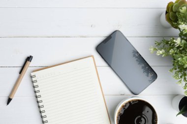 The desk office with a smartphone, blank notepad, coffee cup, and pen on a wood table. Flat lay top view copy space. Concept business technology.