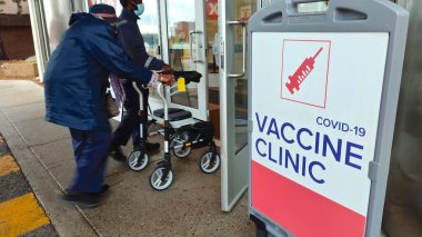 Covid-19 Aşı Kliniği 'nin işareti. Yaşlı bir yetişkinin yürüyüşçüyü iterken görüntüsü bulanık..