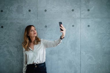 Yeşil gözlü, yetişkin beyaz bir kadın akıllı telefonuyla selfie çekiyor. Ofisinde gri bir duvarın önünde duruyor. Sol elinde siyah bir saat var.. 