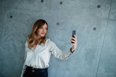 Mutlu, tatmin olmuş yönetici, şirkette teneffüste akıllı telefonunun kamerasına bakıyor. Kafkasyalı yaşlı kadın, modern gri bir duvarın önünde duruyor..