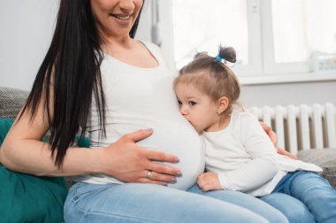 Anne ve kızı kanepede kucaklaşırken at kuyruklu küçük bir kız annesinin koca göbeğini öpüyor. Beyaz saçlı hamile bir kadın sağ eliyle karnını tutuyor. Alyansı nerede?. 