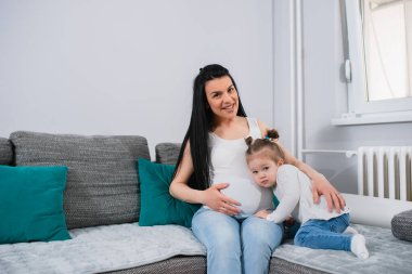 Uzun siyah saçlı, beyaz hamile bir anne kızına sol eliyle sarılıyor ve sağ eliyle karnına sarılıyor. At kuyruklu, küçük sarışın bir kız annemin karnına yaslanıyor..