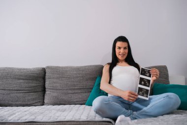 Gelişmiş hamilelikte, siyah, uzun saçlı, beyaz bir kadın yeşil yastıklara yaslanarak oturur. Kocaman göbekli hamile bir kadın, gri bir koltukta bacak bacak üstüne atmış otururken bebeklere ultrason görüntüleri gösteriyor..