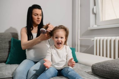Aşırı neşeli bir çocuk mutlu çığlıklar atarken ciddi annesi onun için saç stili yapıyor. Hamile bir anne sağ elinde bir saç fırçası tutar ve sevimli kızını tarar. İki beyaz kadın kanepede oturuyor..