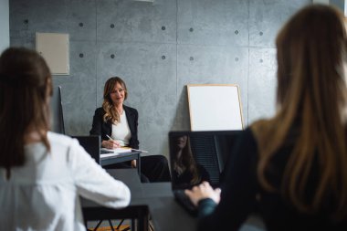 Orta yaşlı beyaz bir öğretim üyesi genç bayan çalışanların bir şirkette beyin fırtınası yapmasını dinlerken not alıyor. İki genç katılımcının arkası dönük. Whiteboard öğretmenin yanında.. 
