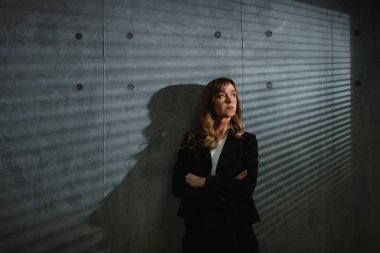 Hayalperest, kahverengi saçlı, resmi giyinmiş beyaz bir kadın koyu renk bir odada kollarını kavuşturarak gri arka planda duruyor. Blinler ve gölgeleri duvarda görünüyor.. 