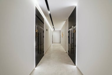 Siyah kapıları ve ışıkları olan beyaz bir koridorun içinde.