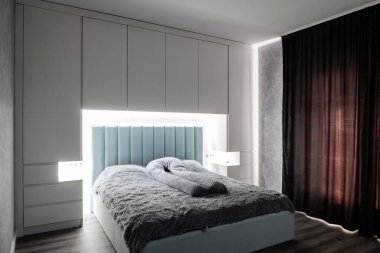 Arka lambalı büyük bir yatak. Penceredeki siyah, koyu perdeler