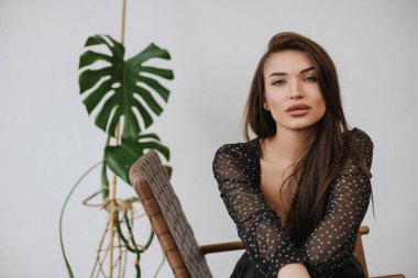 Siyah elbiseli siyah saçlı kız beyaz arka planda bir sandalyede oturuyor.