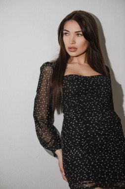 Siyah elbiseli esmer kız stüdyoda beyaz arka planda poz veriyor.