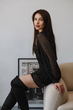 Siyah elbiseli ve yüksek ayakkabılı genç bir kız beyaz bir sandalyede oturuyor.