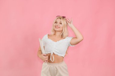 Ellerinde kitapla pembe arka planda poz veren açık renk elbiseli güzel bir kız.