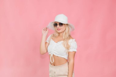 Beyaz şapkalı ve gözlüklü genç bir kız pembe arka planda poz veriyor.