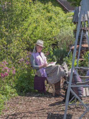 Beervelde, Belçika, 8 Mayıs 2022. Şapkalı, pembe atkılı ve uzun etekli kadın bir sandalyeye oturmuş doğanın ortasında bir kitaba notlar yazıyor.