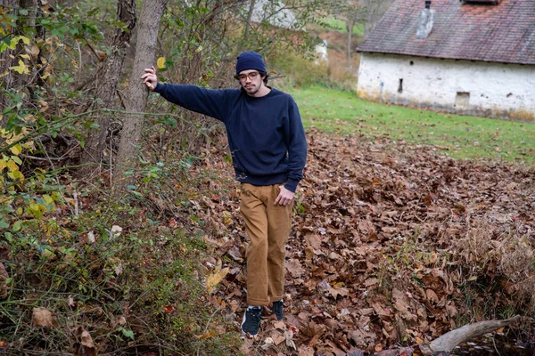 Knappe jongeman leunt op boom op herfstpad door stenen huisje — Stockfoto