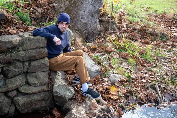 Hombre joven se sienta contra una pared de piedra en el fondo de otoño al aire libre — Foto de Stock