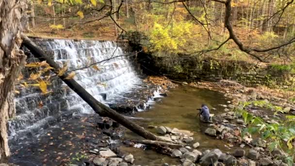 Ein Fotograf am Fuße eines Wasserfalls in farbenfrohen Herbstwäldern — Stockvideo