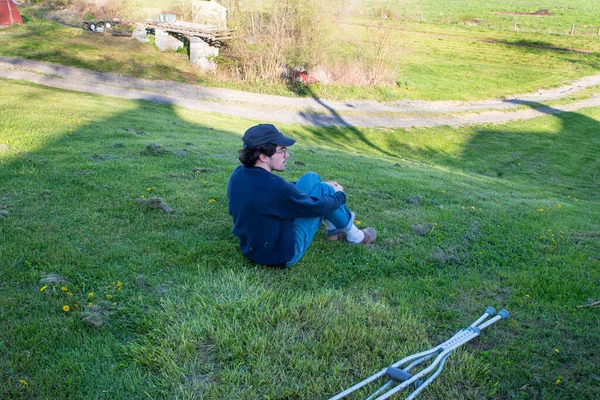 Young man sits alone on grassy rural hill with crutches in foreground — Foto de Stock