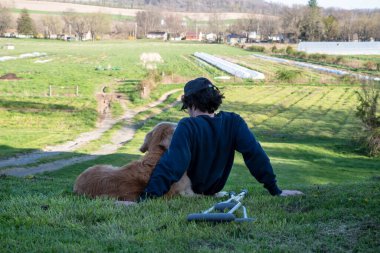 Bu huzurlu kırsal çiftlik arka planının ön planında koltuk değnekleriyle genç bir adam ve sevgili Golden Retriever köpeği kollarını sarıp çimenli bir tepede oturuyorlar..