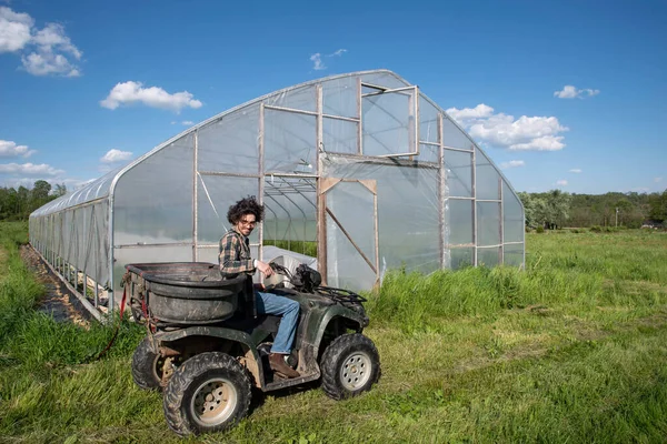 Young farmer on off road vehicle in sunny field by vegetable greenhouse