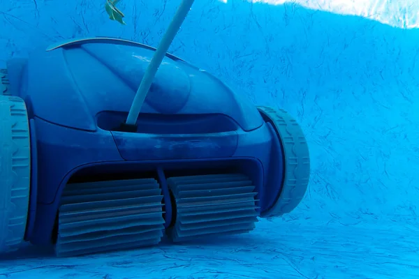 Residential blue vinyl pool liner being cleaned by a motorized wheeled vaccum cleaner with a single leaf in the water. Front wheels and beaters are visible and floating electric cord extends upward