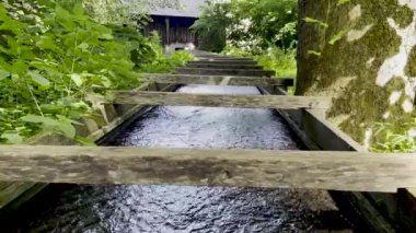 Water flows down a shallow wooden outdoor sluice, into a colonial American iron furnace building in background.Old fashioned hydro electric technology surrounded by nature and green plants