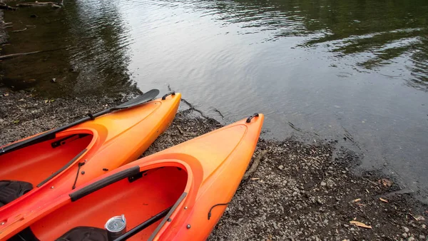 Beautiful outdoor adventure nature background scene with a pair of orange kayaks pointing toward a river and a green woodland scene reflected in the water surface.