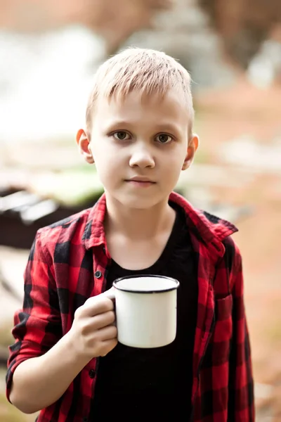 Хлопчик-підліток у картатій сорочці тримає чашку напою. Подорожі, походи, відпустка, концепція кемпінгу . — стокове фото
