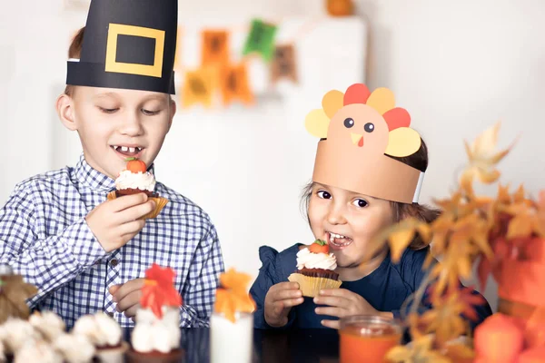 孩子们坐在节日桌上庆祝感恩节.戴着纸制火鸡帽和朝圣者帽的孩子们正在吃纸杯蛋糕和喝牛奶. 图库照片