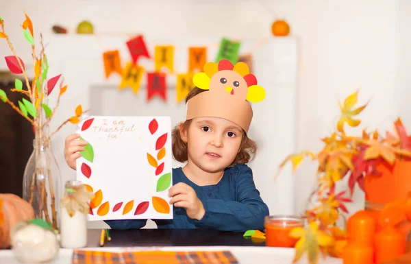 Μικρό χαριτωμένο κορίτσι με χάρτινο καπέλο που γράφει για το οποίο είμαι ευγνώμων. Γιορτάζουμε την ημέρα των Ευχαριστιών. Έργο τέχνης Diy craft. Εικόνα Αρχείου