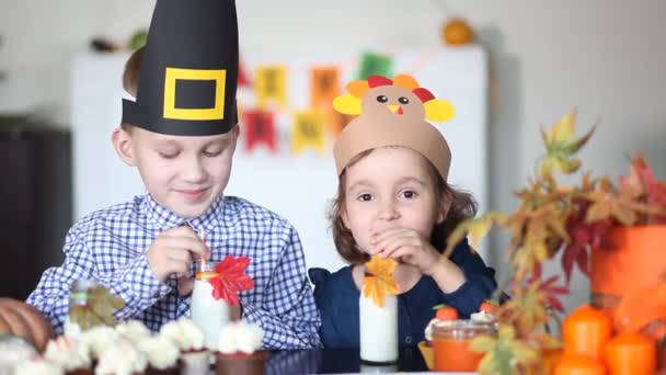 Gyerekek ülnek az ünnepi asztalnál és ünneplik a hálaadást. Papírpulykás sapkás gyerekek és zarándok sapkás gyerekek sütit esznek és tejet isznak. — Stock videók