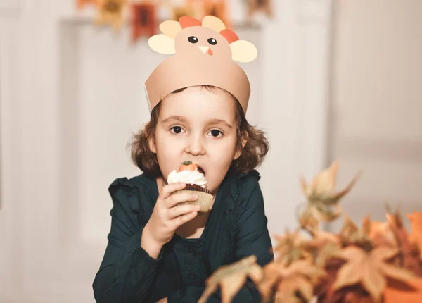 Маленькая девочка в бумажной американской шляпе сидит на праздничном столе и ест кексы. Празднование Дня Благодарения. — стоковое фото