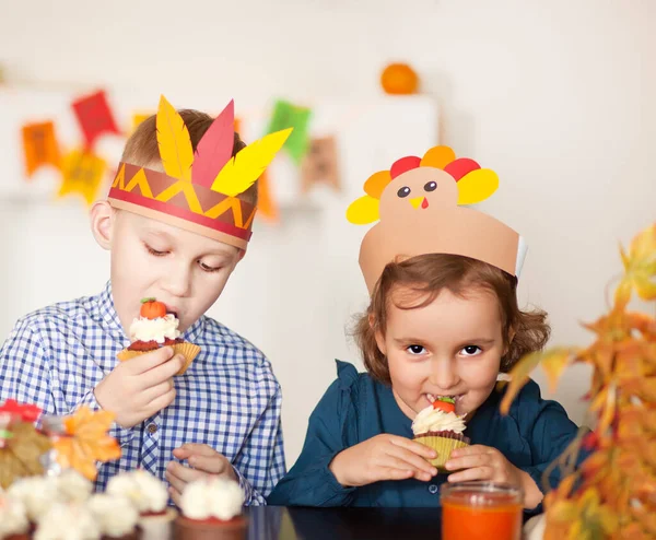 Kinder sitzen auf festlichen Tischen und feiern Erntedank. Kinder mit Papphut und Pilgerhut essen Cupcakes und trinken Milch. lizenzfreie Stockfotos