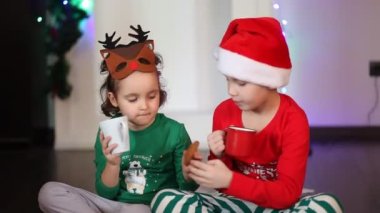 Komik geyik maskeli küçük kız ve Noel Baba şapkalı çocuk kakao içiyor.