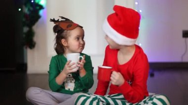 Komik geyik maskeli küçük kız ve Noel Baba şapkalı çocuk kakao içiyor.