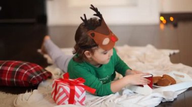 Komik geyik maskesi takan ve Noel pijamalı küçük bir kız ekose bir elbisenin üzerine uzanmış kurabiye yiyor ve kakao içiyor.