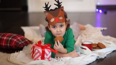 Komik geyik maskesi takan ve Noel pijamalı küçük bir kız ekose bir elbisenin üzerine uzanmış kurabiye yiyor ve kakao içiyor.