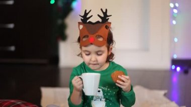 Komik geyik maskesi takan ve Noel pijamalı küçük bir kız. Ekose üzerinde oturmuş kurabiye yiyor ve kakao içiyor.