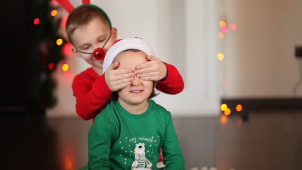 Szczęśliwa mała uśmiechnięta dziewczynka i chłopiec w czapce Mikołaja i maska jelenia siedzi na kocu w piżamie świątecznej. Chłopiec robi niespodziankę dla dziewczyny — Wideo stockowe