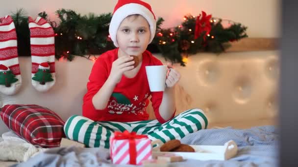 Chłopiec dzieciak w zabawny czerwony Santa czapka i Boże Narodzenie piżama siedzi na łóżko i pije kakao — Wideo stockowe