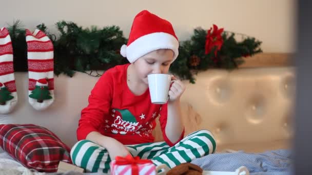 Chłopiec dzieciak w zabawny czerwony Santa czapka i Boże Narodzenie piżama siedzi na łóżko i pije kakao — Wideo stockowe