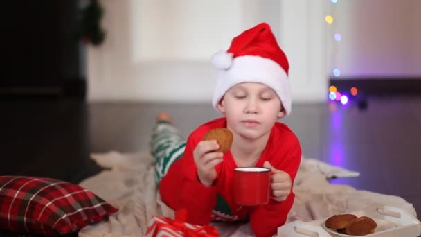 Chłopiec dzieciak w zabawny czerwony Santa czapka i Boże Narodzenie piżama leżą na kratę i picie kakao — Wideo stockowe